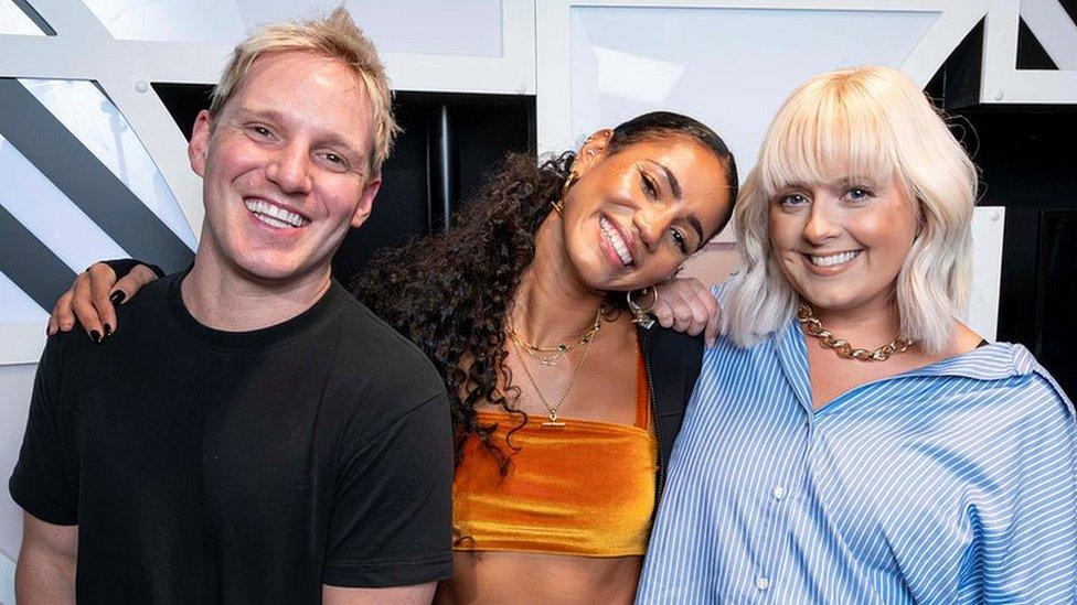 Jamie Laing, Vick Hope and Katie Thistleton in the BBC Radio 1 studio. Jamie is a white man in his 30s with short blonde hair and wears a black T-shirt. Vick is a black woman in her 30s and has her arm around Jamie, her head tilted to the side as she smiles. She has her long hair tired back in a pony tail and wears an orange velvet crop top and black cardigan. Katie is a white woman in her 30s with shoulder length blonde hair. She wears a blue and white striped oversized shirt and smiles at the camera.