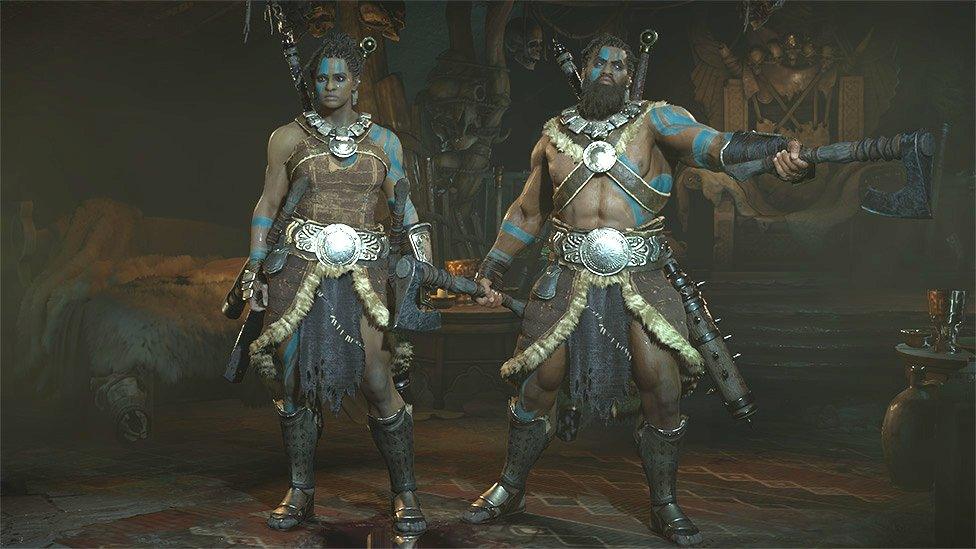 Male and female Barbarian characters from Diablo IV stand side by side. Both are wearing leather armour adorned with large metal buckles and and fur trim. They have darker skin tones, and sport light blue face markings and tribal tattoos in the same colour on their arms. The female character is standing attentively, while the male, who's holding two battleaxes, points one of his weapons menacingly at an unseen foe off-screen