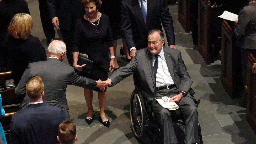 George H W Bush in a wheelchair, reaching for someone's hand, while on his way down a church aisle at his wife's funeral