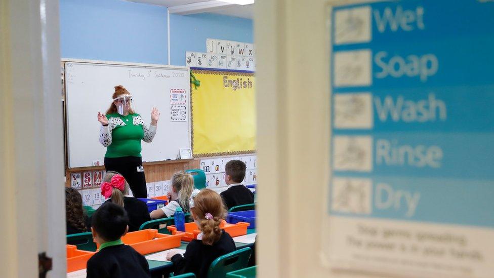 school class, teacher wearing visor and a sign in the forefront saying 'wet, soap,wash,rinse,dry'