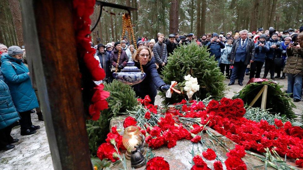 People stand around a mound of wreaths and flowers in Levashovo, with one woman placing a bouquet of white roses on top