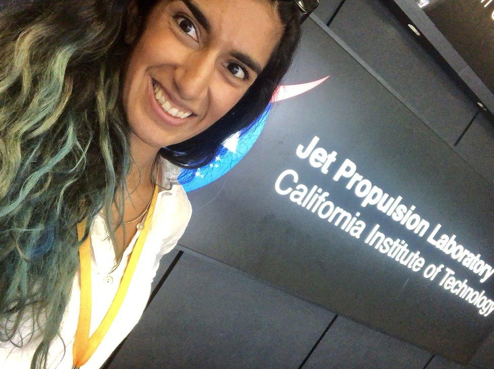 A woman with dark brown and green hair stands in front of a sign reading 'Jet Propulsion Laboratory, California Institute of Technology