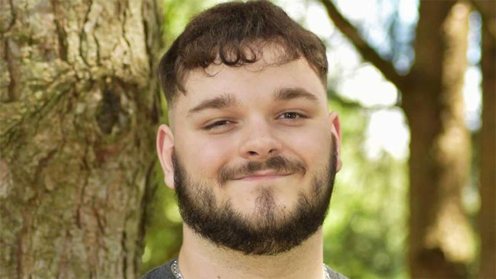 Joe, a white man with a bard, smiling at the camera. The background is of trees and the outdoors, blurred.