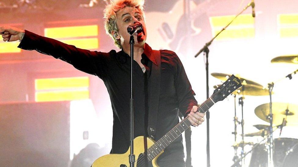 Green Day's Billie Joe holds a guitar with one hand and sings into a microphone during a concert - he's pointing in the air with his free hand