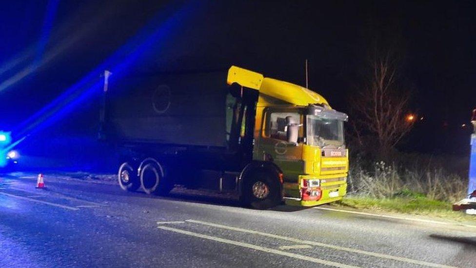 A yellow Scania lorry with blue lights from emergency service cars behind it