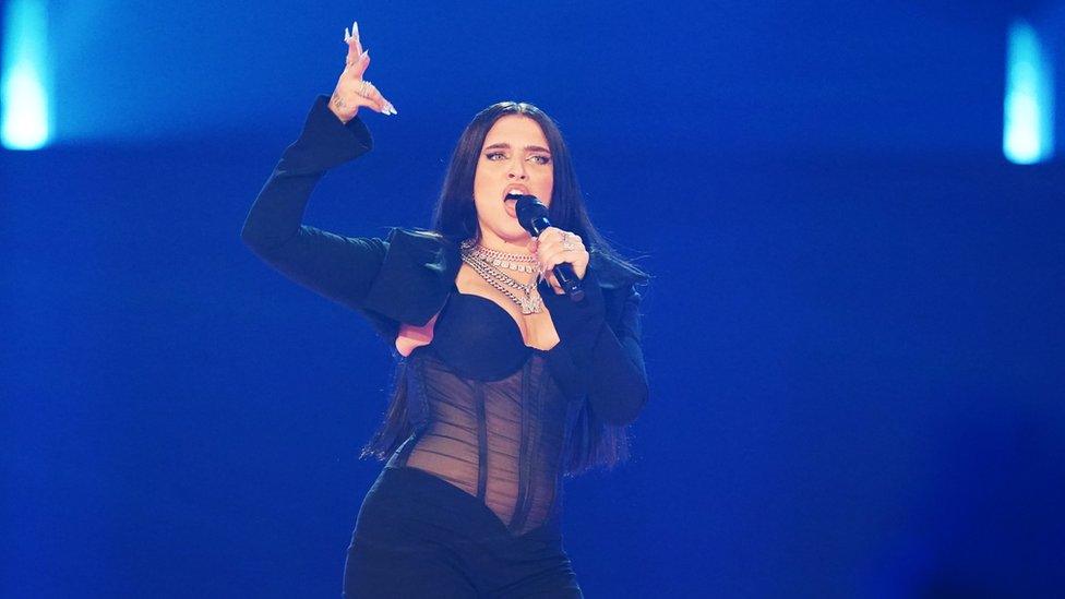 Mae Muller performs on stage at Eurovision. She's wearing a blazer, sheer corset and several chunky silver necklaces. Her hair is down and she's pointing a gun finger in the air.