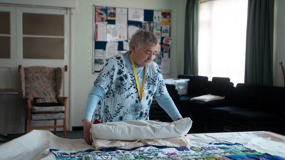 A woman with grey hair and a blue dress working on her large tapestry laid out on a table in a community hall