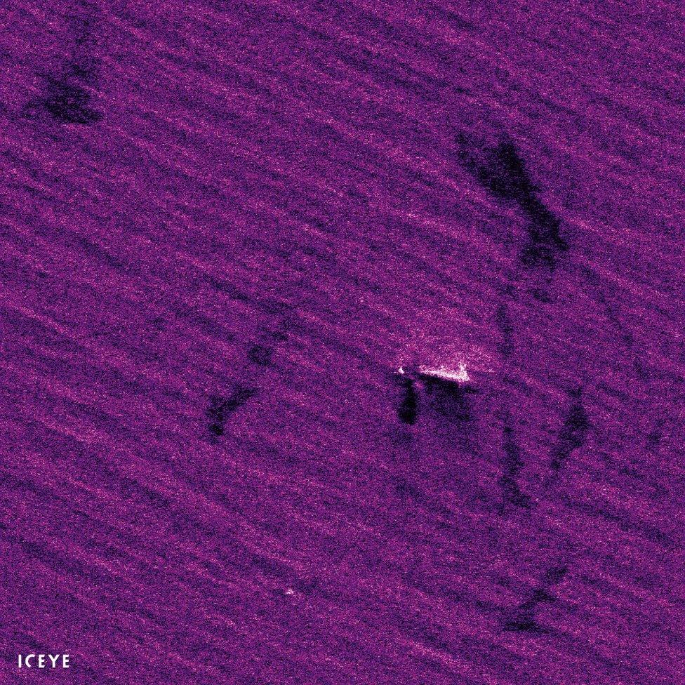 A top-down satellite shot shows a ship upon the ocean, but the colours are unreal and vibrant, with the sea a vivid purple rather than its usual blue - and in this heightened colour state, dark patches can be seen floating on the ocean surface