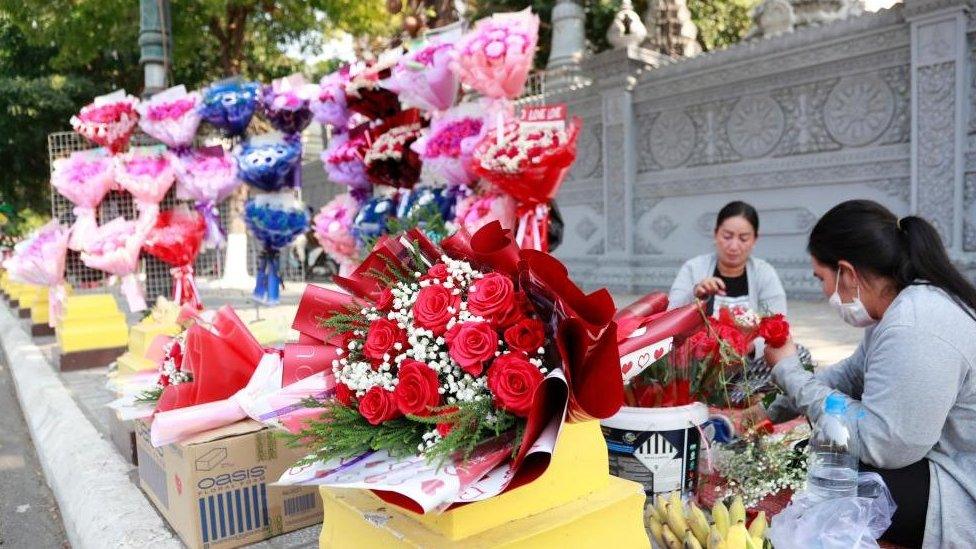 Flower bouquets on display on the side of the street, with women preparing more bunches in the background, in Phnom Penh, Cambodia