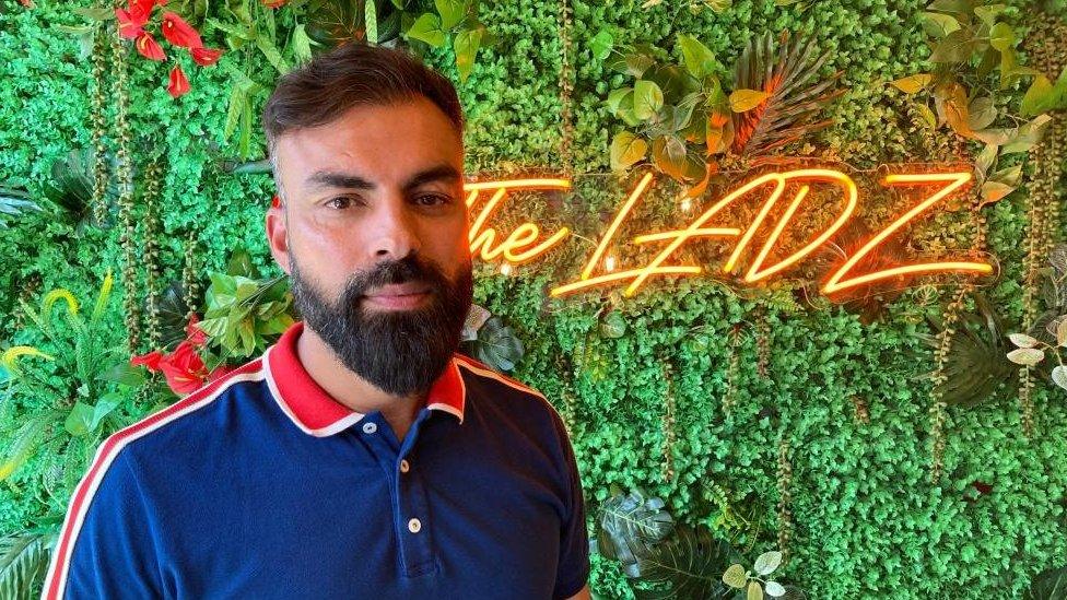 Bearded man stands in front of shrubbery with a sign saying "The Ladz"