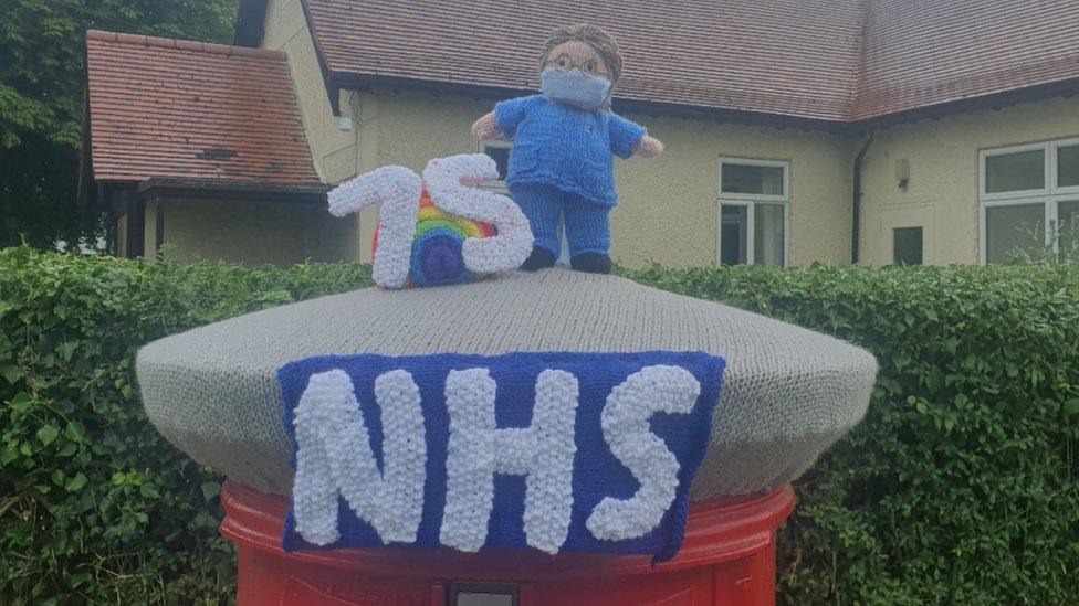 Knitted nurse in uniform wearing a mask stands next to a large "75" on top of a post box