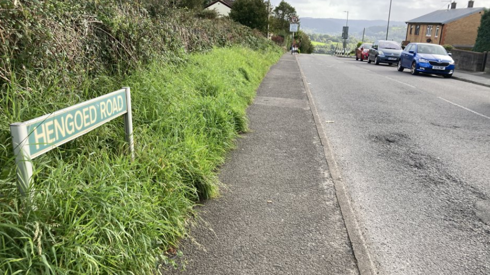 A shot of Hengoed Road, where the incident happened. The road sign is visible as well as a street lined with grass hedges on the left side, and cars and houses on the right side.