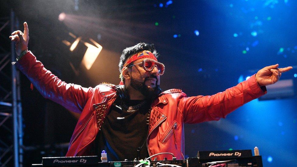 DJ Bobby Friction and the Bhangra All Stars perform on the Truth stage during day 2 of Glastonbury Festival 2023. Bobby is a British Asian man in his early 50s. He wears a studded red leather jacket over a black T-shirt paired with a red headband and red tinted sunglasses. His eyes are closed as he holds both arms out above his DJ deck, his head turned to the left. He's on stage, lit by spotlights and blue light