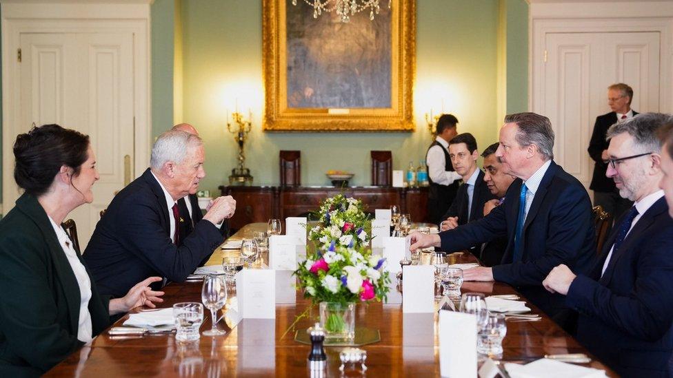 Israeli minister Benny Gantz (second from left) and Lord Cameron (second from right) sit alongside other people around a long wooden table in a chandeliered room