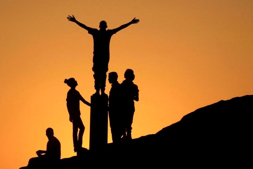 Teenagers stand on top of a rock. Their black silhouettes contrast with the orange sunset.