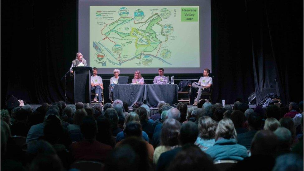 The packed meeting with a panel on stage in front of a graphic of the Heavens landscape