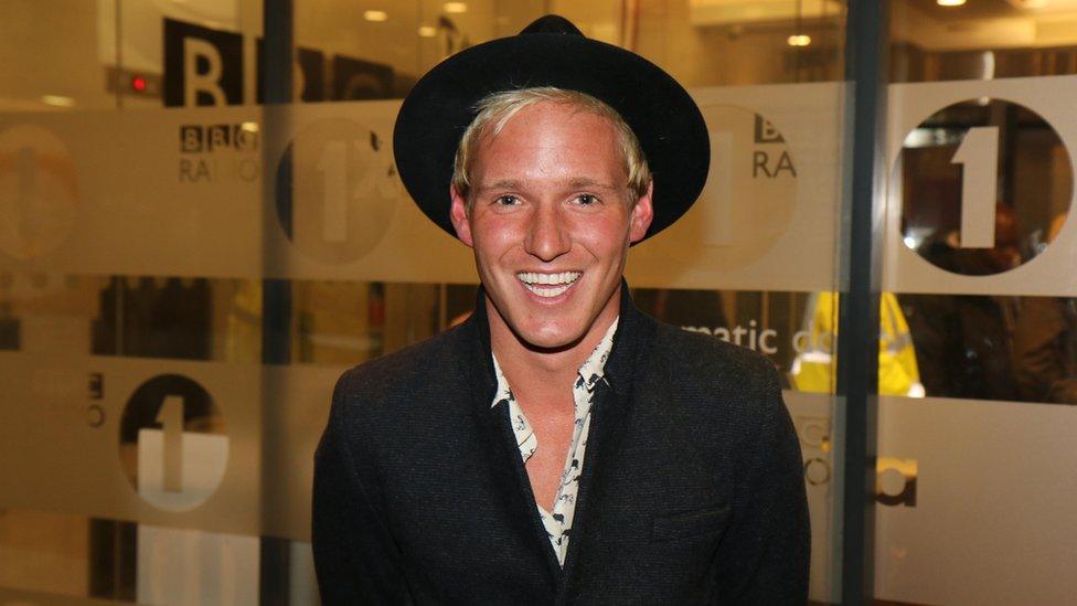 Jamie Laing pictured at BBC Radio 1 offices in 2022. Jamie is a white man with blonde hair in his 30s. He wears a wide-rimmed black hat and a dark coat over a patterned white shirt which is buttoned low. He smiles at the camera and is pictured with BBC Radio 1 branding on windows behind him.