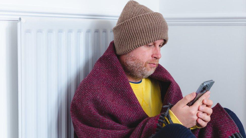 Man in hat and scarf sat next to a radiator