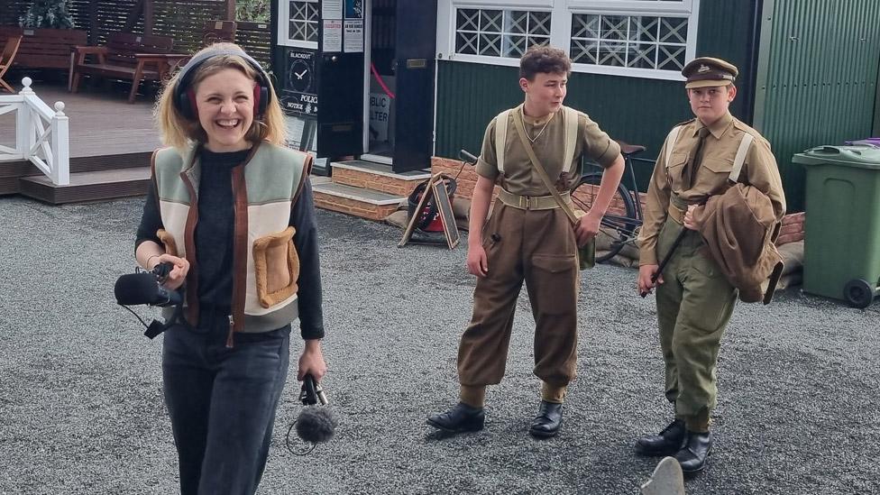 Polly Weston holding a microphone with two young men in WWII uniforms