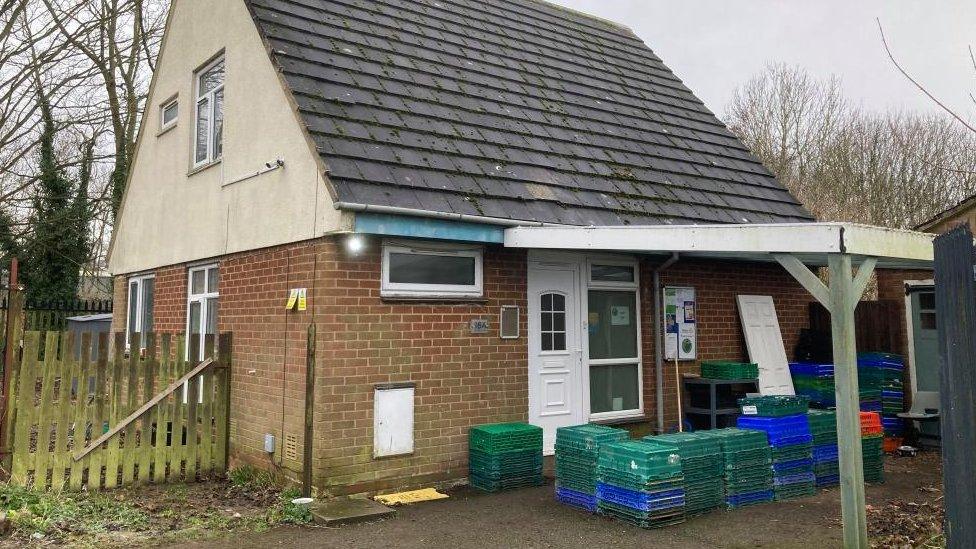 Two-storey brick and render building with pitched roof and wooden car port. Food crates are piled up outside.