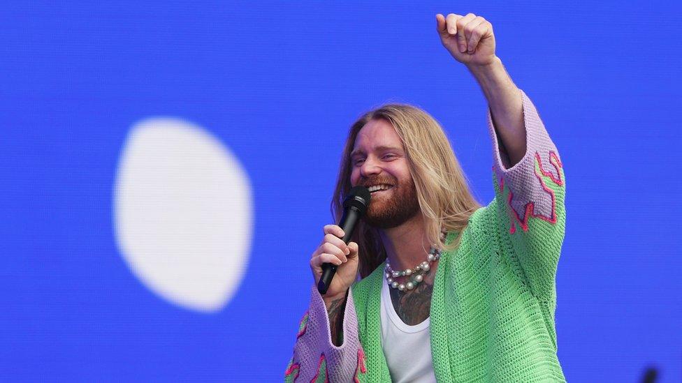 Sam Ryder performs at BBC Radio 2 In The Park 2023. Sam is a white man with long blonde hair and a short strawberry blonde beard. He wears a pink and green knitted cardigan over a white vest and a pearl necklace. He smiles as he sings into a microphone, his right hand held aloft in a fist bump. The staging behind him is blue with a single white circle to his right