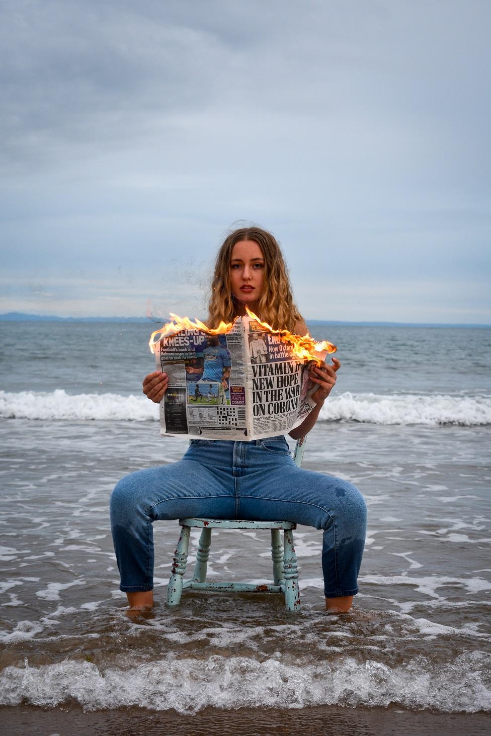 A woman, sits on the beach with water at her feet as she holds a burning newspaper