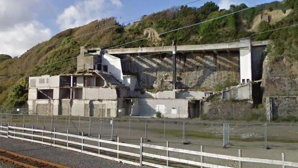 Ruins of the grey concrete building on a cliff face, with fencing in front of it.