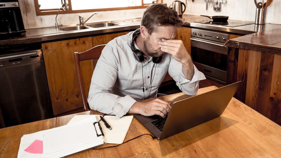 A man rubs his eyes in apparent stress as he sits at a wooden kitchen table with a laptop and papers