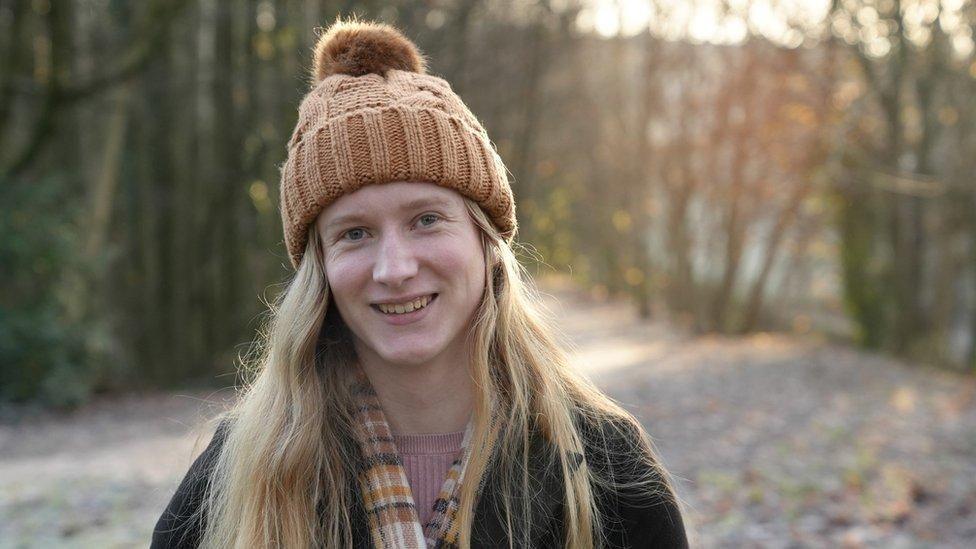 Ellie Gomersall is smiling at the camera. She has long, blonde hair and is wearing a brown, woolly bobble hat, a brown checked scarf and a dark jacket with a pink jumper underneath