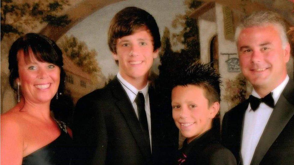 Four people in a photo from left to right. They are looking formally dressed. On the left is June, Jack Thomnas is on the second left and is wearing a suit and black tie, his younger brother Owain stands next to him - who is also smartly dressed and has spikey hair. On the right is the two boy's dad Grant. All are smiling in the photo and they appear to be standing in front of a painting depicting a European country village.