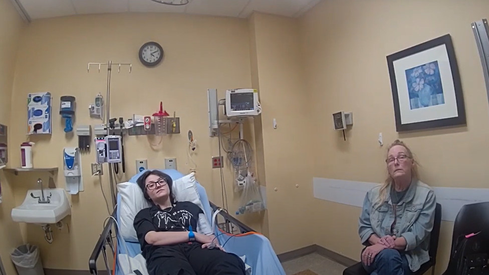 Nex Benedict, who reclines on a hospital bed, speaks to a police officer along with their mother