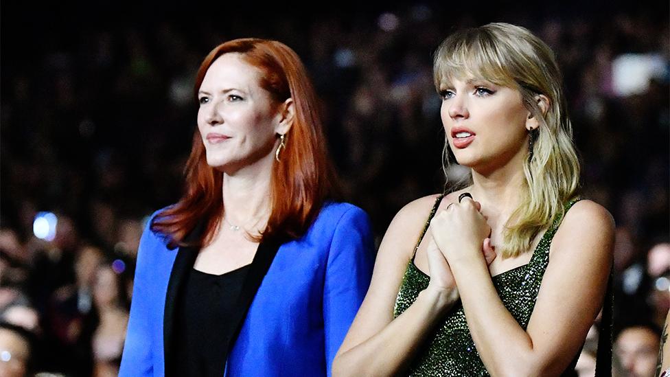 Tree Paine and Taylor Swift, standing together at an awards ceremony. Tree is wearing a blue blazer, and Taylor is wearing a sparkly dress. Taylor looks worried, while Tree has a straight face.