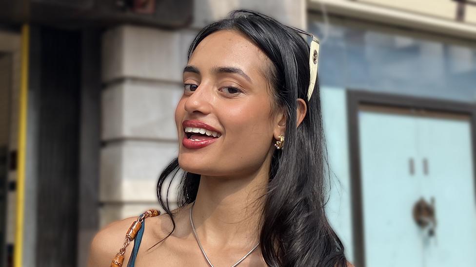 A young Asian woman stands outside a Tube station - the pillars of the entrance visibile in the background. The straps of her blue vest top are visible in this head and shoulders shot. She's smiling, wearing red lipstick and with a pair of sunglasses with thick off-white arms on her head acting as a headband for her long dark hair. She also wears small gold hooped earrings.