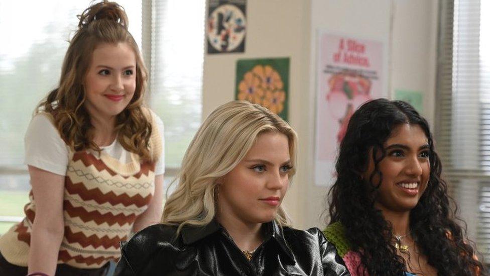Promotional still from 2024 film Mean Girls featuring Bebe Wood as Gretchen Wieners, Reneé Rapp as Regina George and Avantika Vandanapu as Karen Shetty. Gretchen wears her long curly brown hair in a half-up-half-down style, she has brown eyes and wears a brown and cream sweater vest over a white T-shirt. She leans over the table behind Regina, who has long blonde hair flawlessly styled. She wears a black patent style jacket and sits next to Karen who wears her curly dark hair loose. She wears a multi-coloured knitted cardigan. The girls are pictured inside a classroom looking ahead.