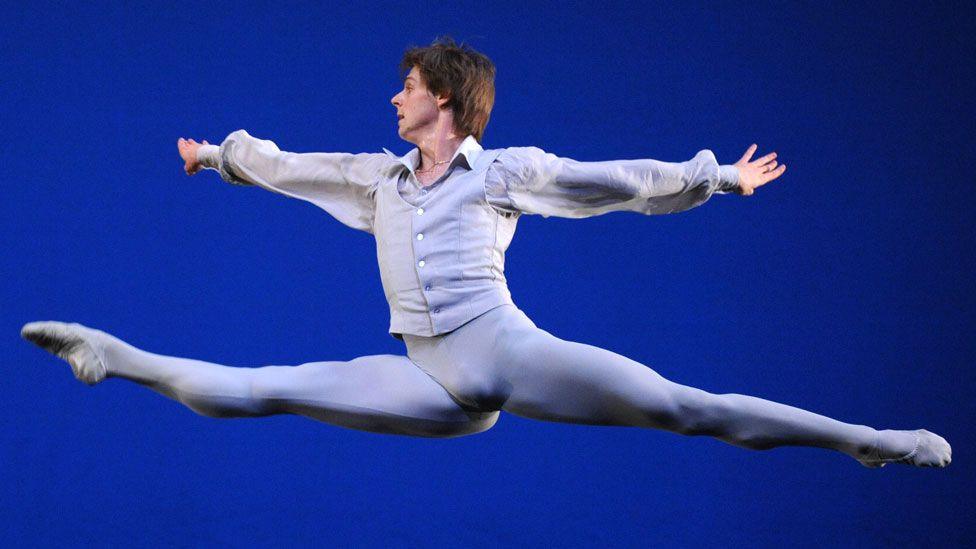 Russian ballet star Vladimir Shklyarov leaps through the air with arms and legs outstretched, wearing a white and grey costume. The background is dark blue.