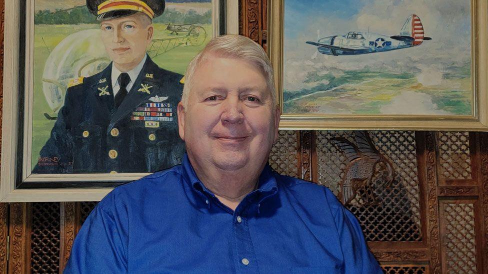 Richard Moseley, sitting in a panelled room with a painted portrait of his father in US Air Force uniform behind him on the left and a painting of an US fighter aeroplane on the right. Richard has short white hair and is wearing a bright blue shirt and starting to smile. 
