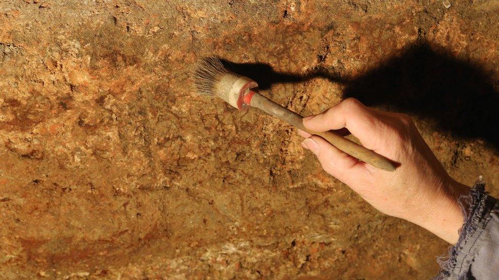 Archaeologist's hand holding a brush against the remains of red paint on the outside Roman wall of the Great Bath