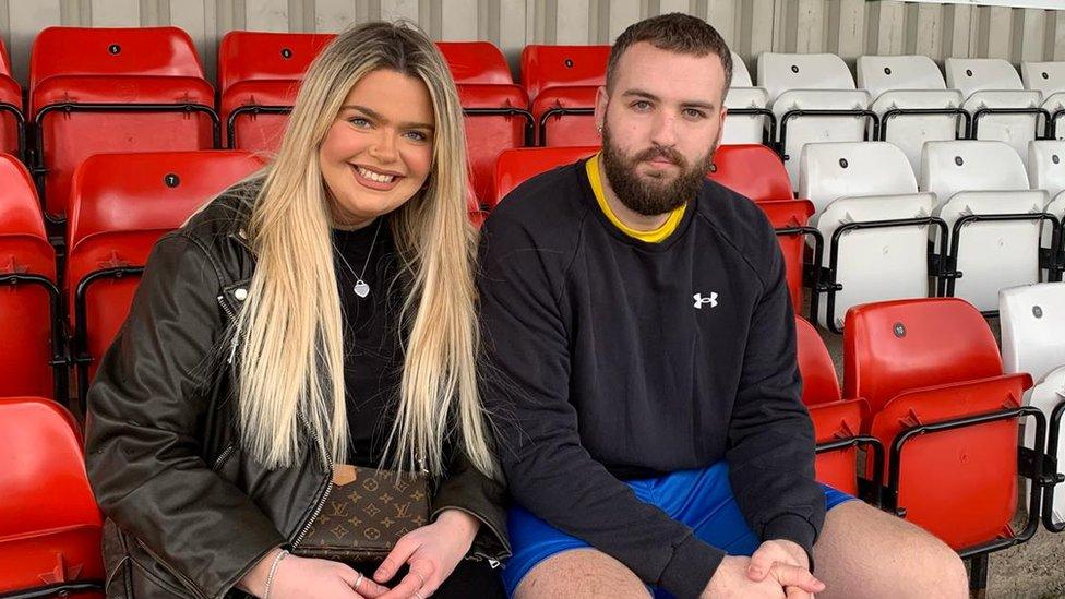 Olivia and Richard Cusack, both sat next to each other in the red and white football stands, and looking at the camera.