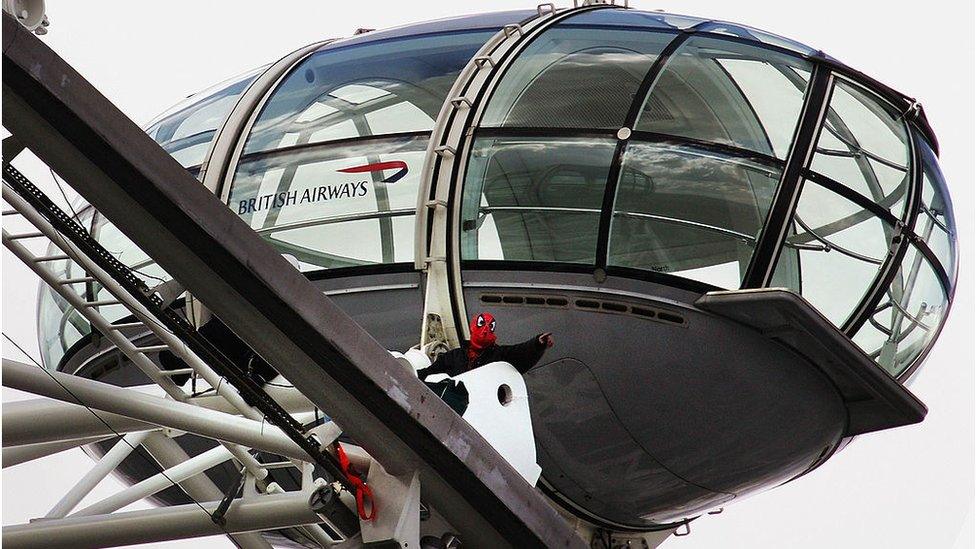 Chick protesting on top of the London Eye wearing a Spider-Man mask and costume under a black coat