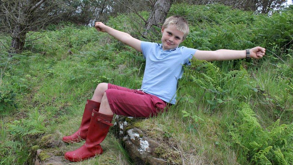 A boy with his arms spread wide sitting on the grass.