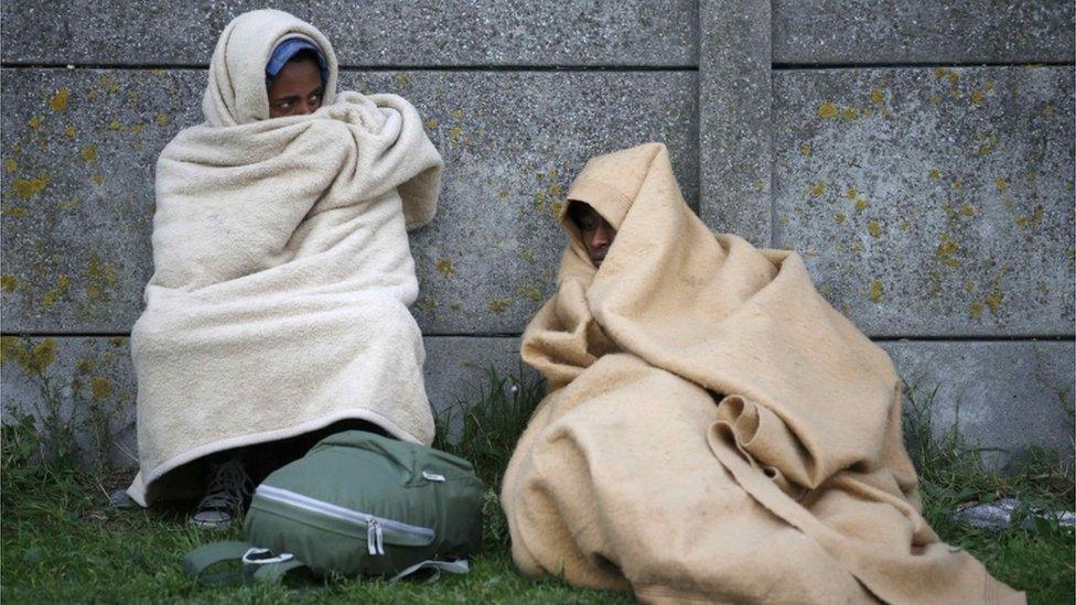 Two migrants cover themselves with blankets and rest against a wall close to the Calais camp.