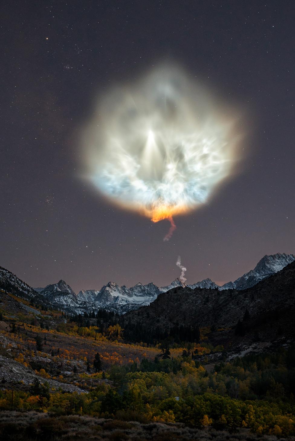 A landscape scene showing mountains at night with a large rocket exhaust plume in the sky