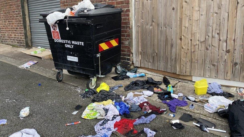 Communal bin with rubbish and clothes on the ground