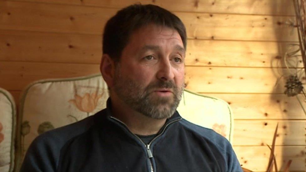 Andy Stallard sitting with a zipped up light jacket in front of a wooden wall