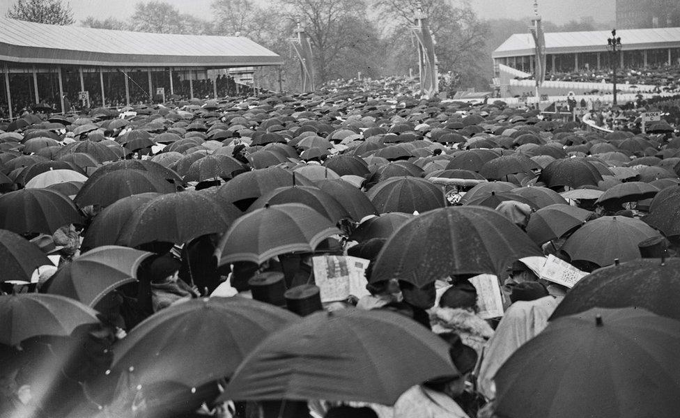 Crowds of well-wishers shelter beneath umbrellas as they wait to catch a glimpse of the Gold State Coach, in which George VI (1895-1952) and Queen Elizabeth (1900-2002) are riding on The Mall