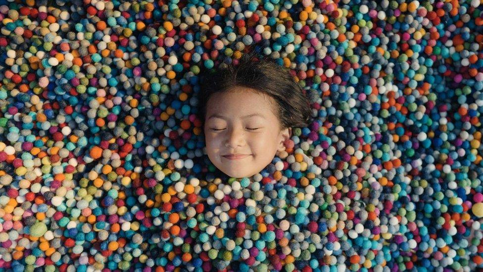 Little girl smiling with her eyes closed surrounded by small colourful felt balls.