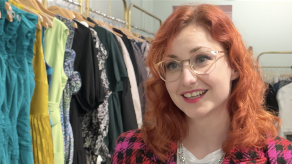 Jess is standing in front of a rail of clothes. She has red shoulder length hair. She is wearing round framed glasses and and a checked pink and black blazer.