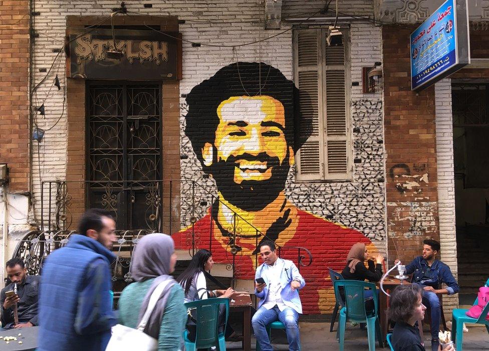 People sitting at a cafe in downtown Cairo, Egypt, with a mural depicting Liverpool FC's Egyptian striker Mohamed Salah painted in the background - 4 April 2018