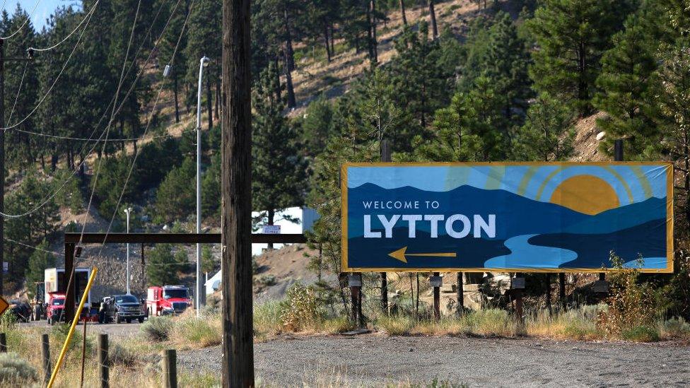 Welcome to Lytton sign is seen as smoke rises over the mountains in Lytton, British Columbia,