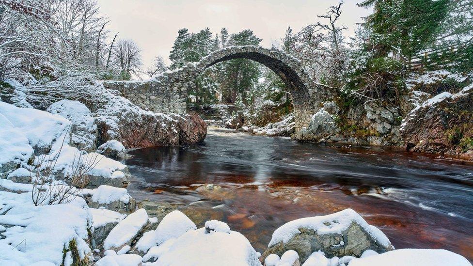 A bridge over a running stream with snow covered rocks and trees around it